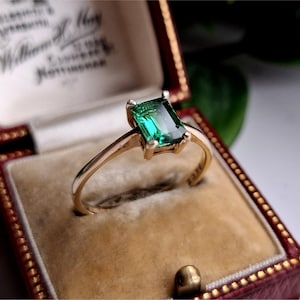 May include: A gold ring featuring a rectangular, emerald-cut green gemstone. The ring is set in a gold band and presented in a red and gold jewellery box. The box has a gold clasp and a cream interior.