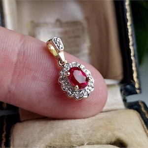 May include: A gold pendant featuring an oval ruby gemstone, encircled by a halo of small diamonds. The pendant has a small bail with diamond detailing. The setting is gold, presenting a classic and elegant design.