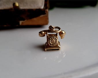 May include: A small, gold-colored charm in the shape of a vintage telephone. The charm features a rotary dial, receiver, and a loop for attaching to a bracelet or necklace. The charm is set against a white surface.