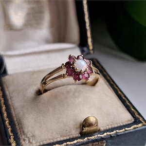 May include: A gold ring featuring a central round white opal surrounded by a cluster of eight deep pinkish-red faceted gemstones arranged in a floral pattern. The band is thin and made of polished gold, showcasing a split shank design.
