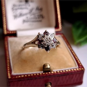 Op de afbeelding: Een vintage gouden ring met een bloemmotief, bezet met kleine, donkerrode edelstenen en heldere accentstenen. De ring wordt gepresenteerd in een rode, met fluweel gevoerde sieradendoos met een gouden sluiting.