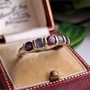 May include: A gold ring featuring five gemstones in alternating purple and burgundy shades. The ring is presented in a red jewellery box with a cream-coloured interior.