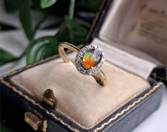 May include: A gold ring featuring an oval-shaped iridescent ammolite gemstone at the center, surrounded by a halo of small clear round stones. The ring has a smooth, polished gold band and is presented inside a dark blue velvet-lined jewelry box.