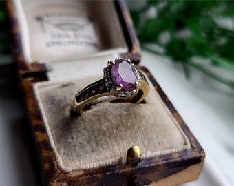 Puede incluir: Un anillo de oro con una gran piedra preciosa ovalada de color púrpura rodeada de diamantes más pequeños. El anillo está en una caja de anillo de terciopelo marrón.