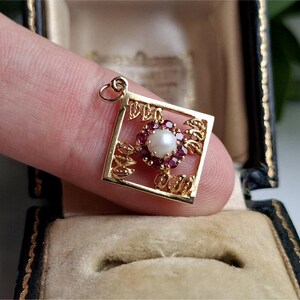 May include: A gold pendant with a square frame. It features a central pearl surrounded by small red gemstones. The pendant has a floral design.