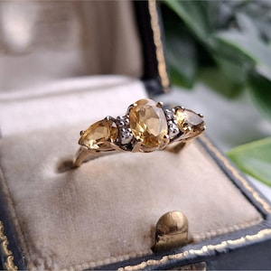 May include: A gold ring featuring a central oval-cut yellow citrine flanked by two smaller pear-cut citrines, accented with small clear stones set in the metal band, presented inside a dark blue velvet-lined jewelry box.