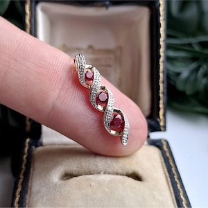 May include: A gold pendant featuring a twisted, ribbon-like design set with three round, deep red rubies and accented with small, sparkling white diamonds along the edges of the metal curves, creating a vertical, elegant jewelry piece.