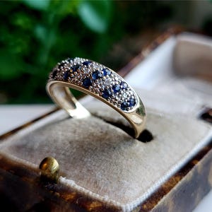 May include: A gold band ring set with a row of blue and white gemstones. The ring is displayed in a velvet-lined box.