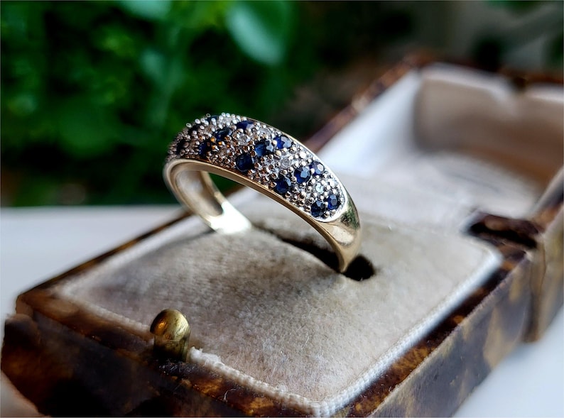 May include: A gold band ring set with a row of blue and white gemstones. The ring is displayed in a velvet-lined box.