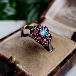 May include: A silver ring featuring a cluster of pink gemstones surrounding a central opal. The ring is presented in a brown velvet ring box.