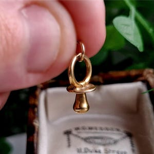 Peut inclure: Un pendentif en forme de sucette pour bébé en or. Le charme a la forme d'une sucette traditionnelle avec un dessus rond et une petite poignée.