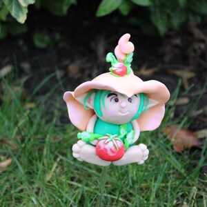 Könnte beinhalten: Eine kleine, fantasievolle Figur eines Zwergs, der einen pfirsichfarbenen Hut mit einem grünen Blatt oben trägt. Der Zwerg hält eine rote Tomate in seinen Händen und hat grünes Haar und ein hellrosa Gesicht.