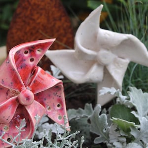 Könnte beinhalten: Zwei Keramik-Windrad-Skulpturen. Eine ist leuchtend rot mit dunkelrosa Akzenten und kleinen Löchern, die andere ist weiß. Sie sind inmitten von Grün angeordnet und schaffen eine gartenähnliche Szene.