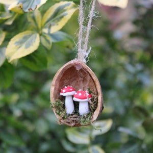Könnte beinhalten: Ein skurriles Ornament mit zwei Miniatur-Pilzfiguren in Rot und Weiß, die in einer Walnussschale liegen. Die Schale wird von einer Schnur gehalten, mit grünem Moos. Der Hintergrund ist ein verschwommener Blick auf grünes Laub.