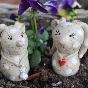 Könnte beinhalten: Zwei Keramik-Hasenfiguren mit weißen Punkten, eine mit einem roten Herzen und die andere mit einem silbernen Anhänger. Die Hasen sitzen auf einem Bett aus braunem Mulch.