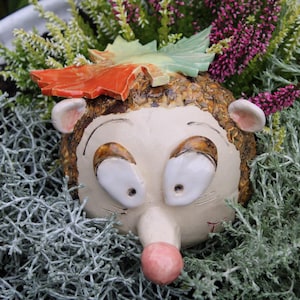 Könnte beinhalten: Eine Igel-Figur aus Keramik mit cremefarbenem Gesicht, großen weißen Augen, einer rosa Nase und einem strukturierten braunen Rücken. Ein dekoratives orange-grünes Keramikblatt ist oben am Kopf des Igels befestigt und setzt einen farbigen Akzent.
