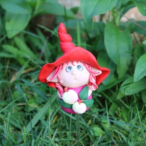 Könnte beinhalten: Eine kleine rote Gnomfigur mit einem spitzen Hut und einem rosa Kleid, die weiße Blumen hält. Der Gnom steht in einem Bett aus grünem Gras.