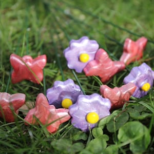 Könnte beinhalten: Eine Sammlung dekorativer blütenförmiger Gartenstecker in Rot- und Lilatönen mit gelben Zentren. Die Stecker sind in grünem Gras angeordnet und schaffen einen farbenfrohen und skurrilen Gartenakzent.