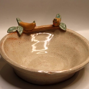 Könnte beinhalten: Eine Keramikschale mit gesprenkelter beigefarbener Oberfläche. Zwei kleine, orangefarbene Vögel mit grünen Blättern sitzen auf einem braunen Ast, der sich über den Rand der Schale wölbt. Die Schale ist rund und flach.
