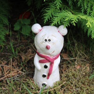 Könnte beinhalten: Eine weiße Keramikfigur in Form eines Schneemanns mit glänzender Oberfläche. Die Figur hat einen rosa bemalten Schal, eine kleine rosa Nase und drei schwarze, runde Knöpfe auf der Vorderseite. Sie besitzt zwei runde Ohren und ein Gesicht.