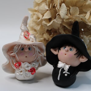 Könnte beinhalten: Zwei skurrile Figuren, eine in weißem Hut und Kleid mit roten Akzenten, die andere in schwarzem Anzug und Hut. Beide haben überraschte Ausdrücke und stehen vor einem Hintergrund aus getrockneten Blumen. Die Figuren sind wahrscheinlich handgefertigt.