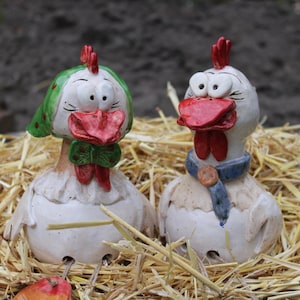 Könnte beinhalten: Zwei skurrile Keramik-Hühnerfiguren. Eines trägt einen grünen Polka-Dot-Hut und eine Fliege, das andere einen blauen Schal. Beide haben rote Kämme und Schnäbel, die auf einem Strohbett liegen. Ein bezaubernder Dekorationsartikel.