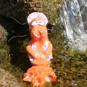 Könnte beinhalten: Orangefarbene Keramikfigur einer Kreatur mit großen Augen, einem rosa-weißen Kopfschmuck und Drahtbart. Die Figur befindet sich in einem Bach, umgeben von Felsen und Moos.