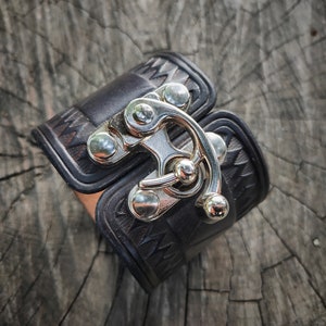 May include: A black leather cuff bracelet with a silver metal clasp. The clasp has a decorative design and is secured with a small silver screw.