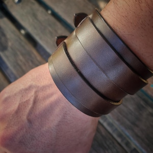 May include: A brown leather cuff bracelet with several stacked bands. The bracelet is a dark brown colour and appears to be made of genuine leather. The cuff is worn on the wrist and has a rugged, masculine aesthetic.