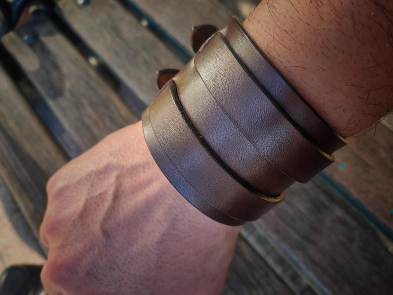May include: A brown leather cuff bracelet with multiple stacked bands. The bracelet is a rich, dark brown color and appears to be made of genuine leather. The cuff is worn on the wrist and has a rugged, masculine aesthetic.