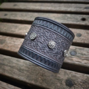 May include: A dark gray cuff bracelet with a textured surface and silver-colored button accents. The bracelet has a geometric pattern along the top and bottom edges. The cuff is approximately 2 inches wide.