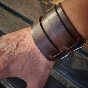 May include: A wide, brown leather cuff bracelet with a buckle closure. The bracelet is made of several thick, overlapping straps of leather. The metal buckle is silver in color. The bracelet is worn on a wrist.