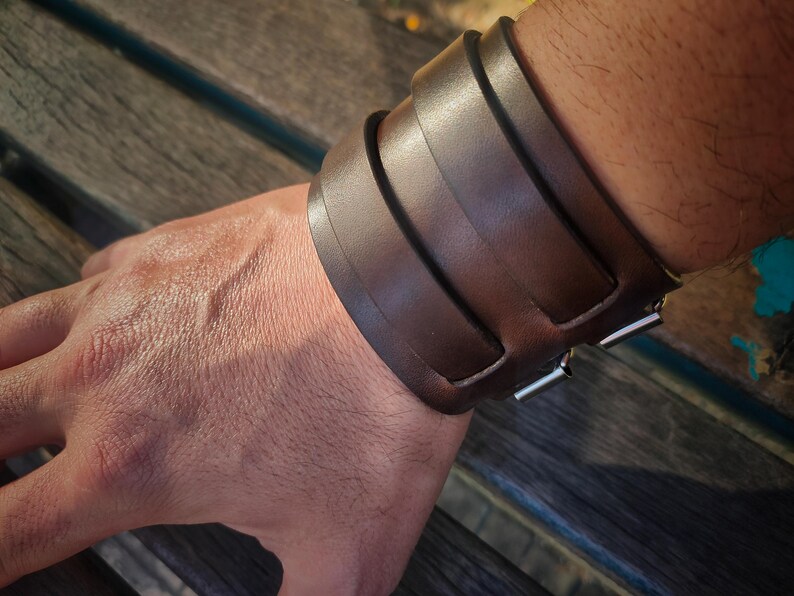 May include: A wide, brown leather cuff bracelet with a buckle closure. The bracelet is made of several thick, overlapping straps of leather. The metal buckle is silver in color. The bracelet is worn on a wrist.
