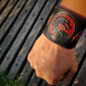 Pode incluir: Pulseira de couro preto com um design de dragão vermelho e preto. A pulseira tem costuras douradas e um fundo vermelho.