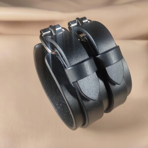 May include: Black leather wrist cuffs with silver-tone buckles. The cuffs are stacked and feature adjustable straps. The leather appears smooth and the overall design is minimalist and modern.