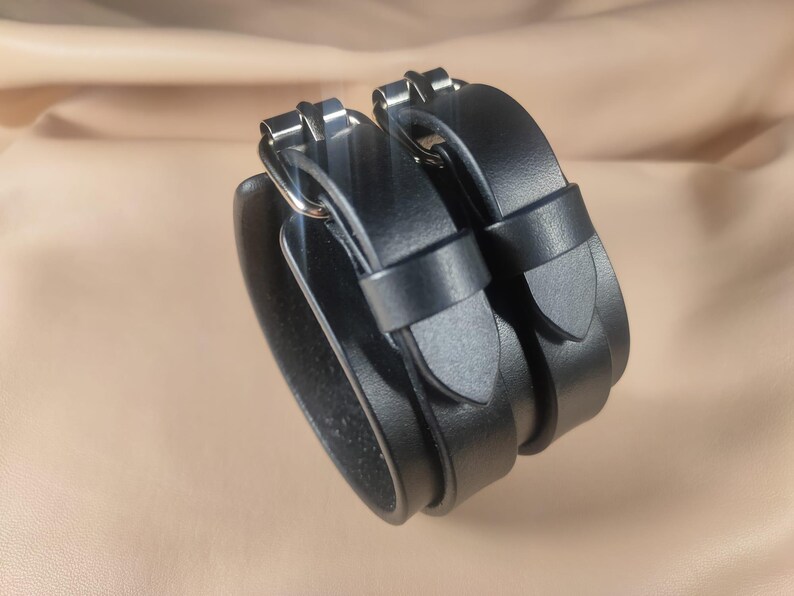 May include: Black leather wrist cuffs with silver-tone buckles. The cuffs are stacked and feature adjustable straps. The leather appears smooth and the overall design is minimalist and modern.