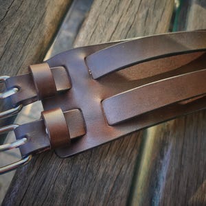 May include: Close-up of a brown leather belt with two silver-colored buckles and two matching leather loops. The belt has two parallel leather handles. The leather has a smooth texture and a rich, warm brown color.