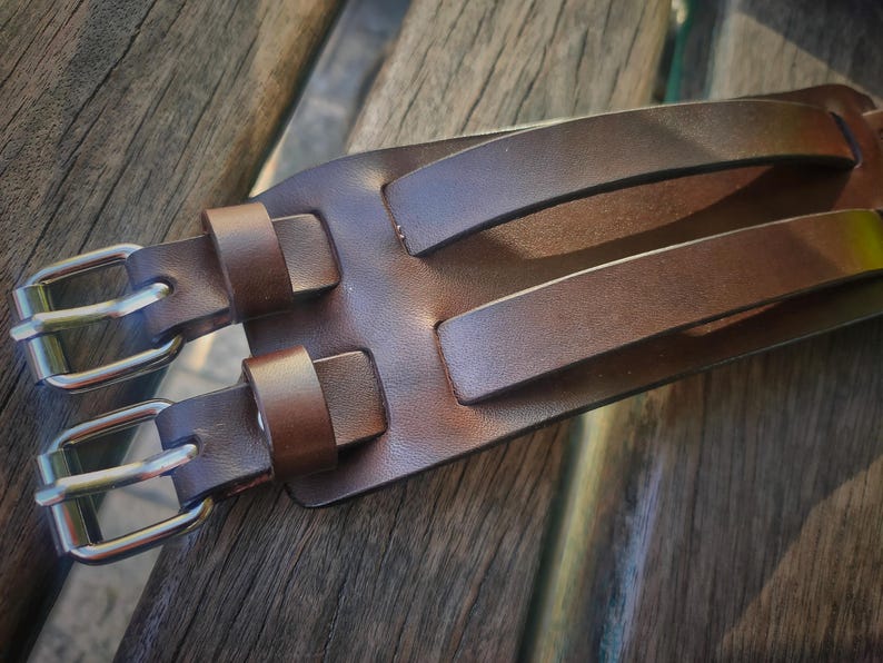 May include: Close-up of a brown leather belt with two silver-colored buckles and two matching leather loops. The belt has two parallel leather handles. The leather has a smooth texture and a rich, warm brown color.