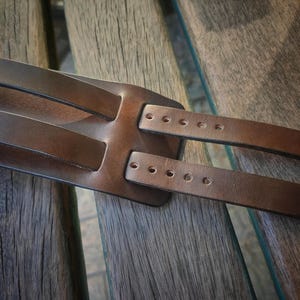 May include: Close-up of a brown leather guitar strap. The strap has a wide base with two narrower straps extending from it. Each strap has several holes for adjustment. The leather appears to be well-worn and has a natural grain.