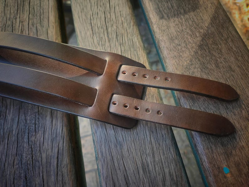 May include: Close-up of a brown leather guitar strap. The strap has a wide base with two narrower straps extending from it. Each strap has several holes for adjustment. The leather appears to be well-worn and has a natural grain.