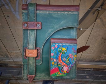 May include: A teal green leather notebook with a brown leather flap and a brown leather strap. The notebook has a smaller pocket on the front with a colorful illustration of a cat in a garden.