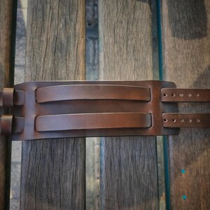 May include: A brown leather wrist cuff with two straps and silver buckles. The cuff has two raised sections and is designed to wrap around the wrist. The leather has a rich, textured appearance.