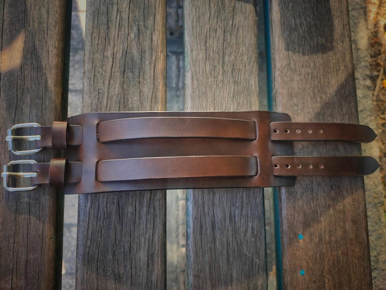 May include: A brown leather wrist cuff with two straps and silver buckles. The cuff has two raised sections and is designed to wrap around the wrist. The leather has a rich, textured appearance.