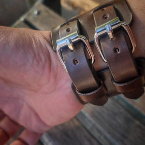May include: A brown leather wrist cuff with two silver buckles. The cuff is made of thick leather and appears to be handmade. The buckles are shiny and add a touch of elegance to the rugged design.