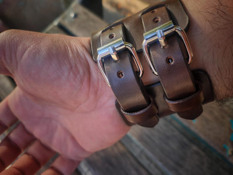 May include: A brown leather wrist cuff with two silver buckles. The cuff is made of thick leather and appears to be handmade. The buckles are shiny and add a touch of elegance to the rugged design.