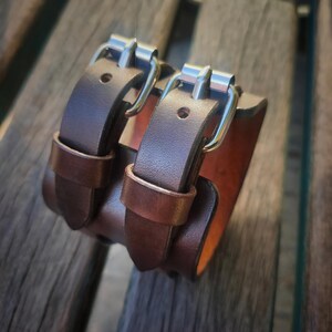 May include: A brown leather wrist cuff with two straps and silver-colored buckles. The cuff is made of thick leather and has a contrasting lighter brown interior. The cuff is designed to be worn around the wrist.
