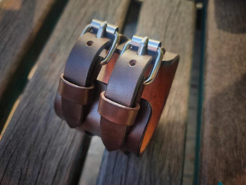 May include: A brown leather wrist cuff with two straps and silver-colored buckles. The cuff is made of thick leather and has a contrasting lighter brown interior. The cuff is designed to be worn around the wrist.