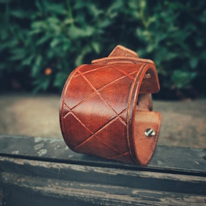 May include: A brown leather cuff bracelet with a diamond pattern. The bracelet has a buckle closure and is made of high-quality leather.