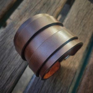 May include: A brown leather cuff bracelet with a layered design. The bracelet appears to be made of several strips of leather stacked on top of each other. The cuff is resting on a wooden surface.