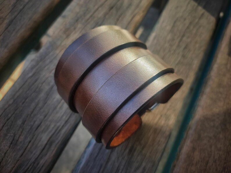 May include: A brown leather cuff bracelet with a layered design. The bracelet appears to be made of several strips of leather stacked on top of each other. The cuff is resting on a wooden surface.